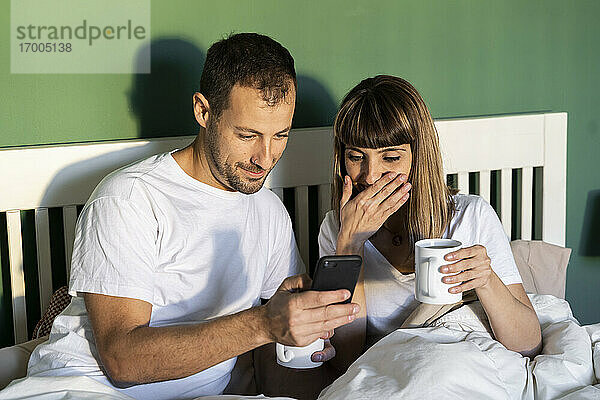Freund und Freundin mit Kaffeetasse und Mobiltelefon  während sie zu Hause im Bett sitzen
