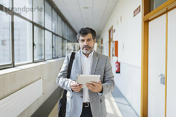 Reifer männlicher Professor  der ein digitales Tablet in einem Korridor einer Universität hält