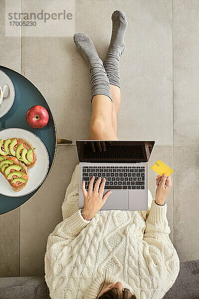 Frau kauft online mit Kreditkarte über Laptop ein  während sie zu Hause auf dem Boden sitzt