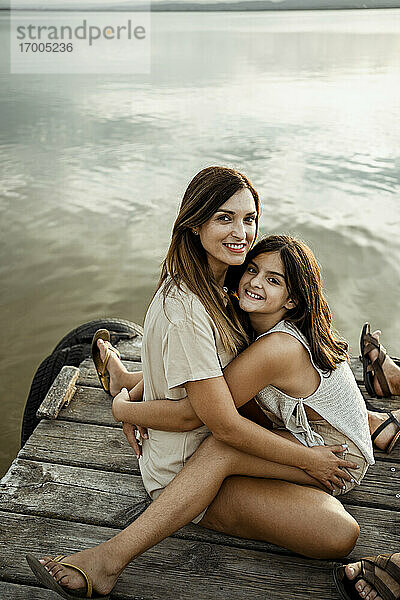 Mutter und Tochter umarmen sich  während sie am Steg am See sitzen