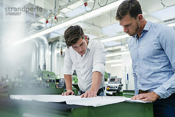 Männlicher Unternehmer mit einem Kollegen  der eine Blaupause auf dem Schreibtisch in einer Fabrik betrachtet