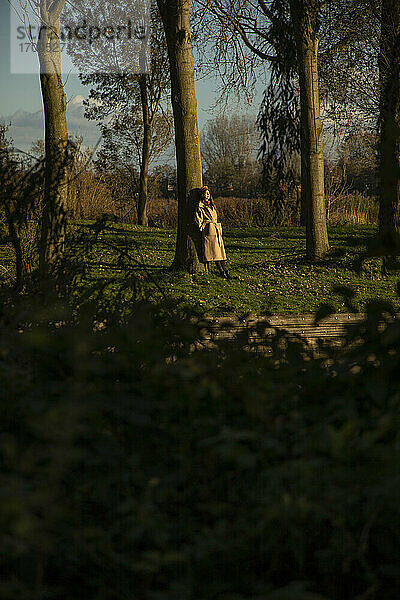 Mittlere erwachsene Frau lehnt an einem Baumstamm in einem öffentlichen Park im Herbst