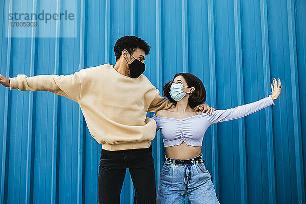 Unbekümmertes Paar mit Gesichtsmaske tanzt an einer blauen Wand