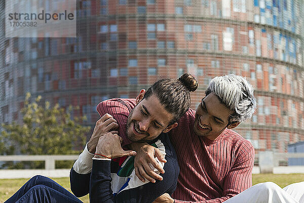 Lächelnder Mann  der sich umarmt  während er seinen männlichen Partner anschaut  während er im Park sitzt