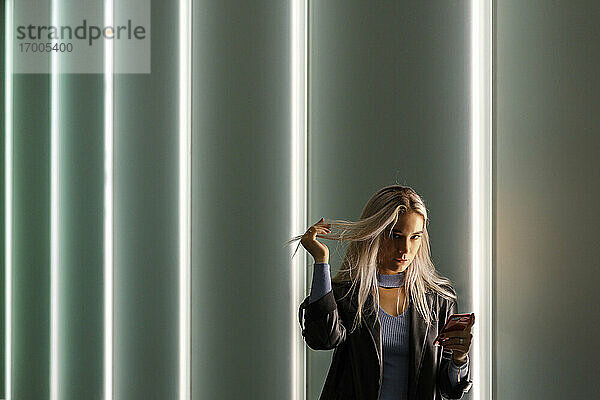 Attraktive Frau hält Haare in der Hand mit Smartphone  während sie vor einer beleuchteten Wand steht