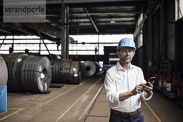Geschäftsmann hält Smartphone in der Hand  während er in einer Fabrik steht