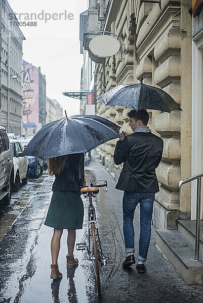 Männliche und weibliche Kollegen  die während der Regenzeit mit dem Fahrrad auf dem Bürgersteig spazieren gehen