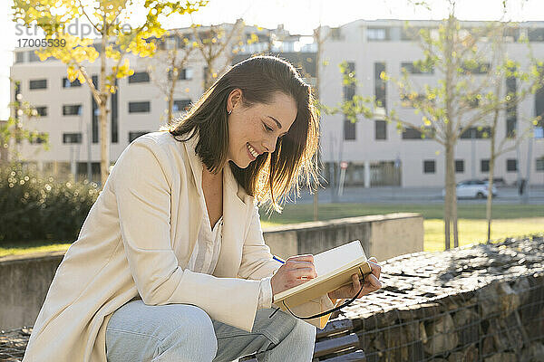 Lächelnde Geschäftsfrau  die auf einer Mauer sitzend in ihr Tagebuch schreibt