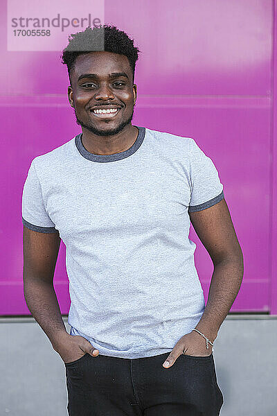 Lächelnder junger Mann mit Händen in den Taschen  der an einer rosa Wand steht