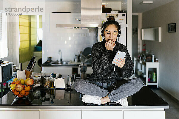 Teenager-Mädchen mit Kopfhörern  das Obst isst  während es ein Handy benutzt und zu Hause sitzt