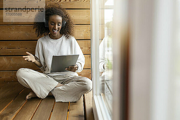 Junge Frau mit Kopfhörern  die ein Videogespräch über ein digitales Tablet führt  während sie zu Hause am Fenster sitzt