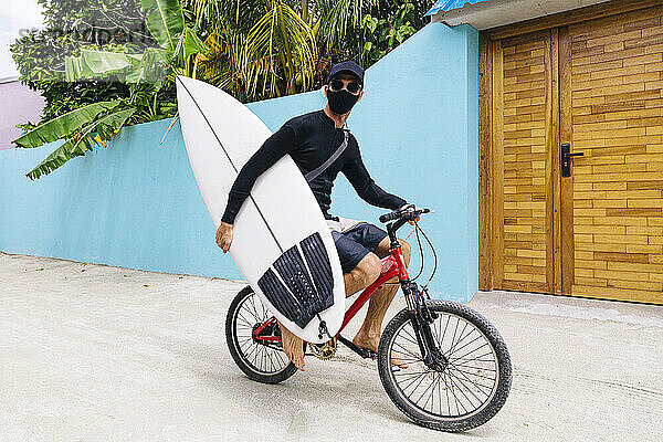 Mann mit Gesichtsschutz  der ein Surfbrett trägt  während er auf dem Fußweg auf der Insel Huraa  Malediven  Fahrrad fährt