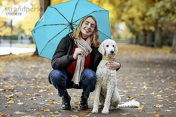 Lächelndes Teenager-Mädchen  das einen Regenschirm hält  während es neben seinem Haustier auf der Straße im Park hockt