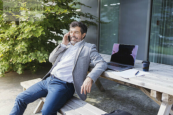 Lächelnder Geschäftsmann  der über sein Smartphone spricht und sich auf einen Tisch vor einem Bürogebäude lehnt
