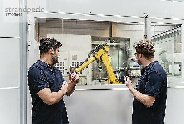 Männliche Mitarbeiter betrachten einen Roboterarm durch ein Fenster  während sie in einer Fabrik stehen