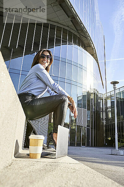 Lächelnde Geschäftsfrau sitzt auf einer Bank neben einem Bürogebäude