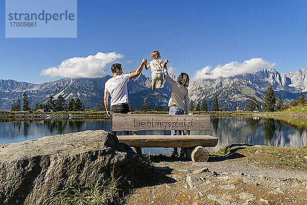 Familie mit einer kleinen Tochter steht gemeinsam hinter einer Seeuferbank im Kaisergebirge