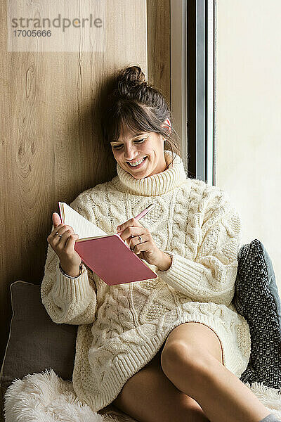 Lächelnde Frau schreibt in ein Buch  während sie zu Hause am Fenster sitzt