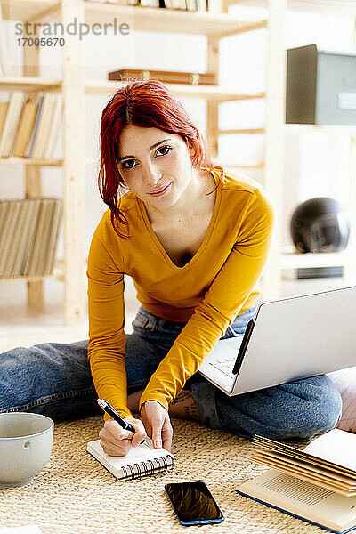 Junge Frau mit Laptop auf dem Schoß schreibt in Notizblock beim Lernen zu Hause