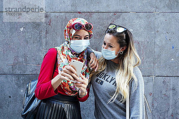 Muslimische Frau teilt Smartphone mit Freundin vor grauer Wand