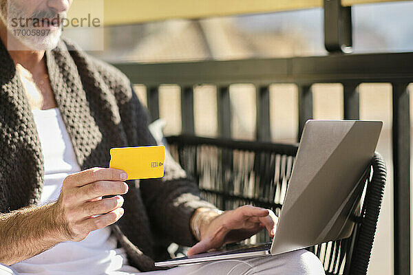 Älterer Mann zu Hause mit Kreditkarte beim Online-Shopping auf dem Laptop an einem sonnigen Tag