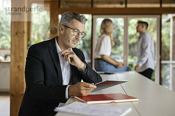 Geschäftsmann  der ein digitales Tablet benutzt  während er zu Hause an einem Tisch mit Kollegen im Hintergrund steht