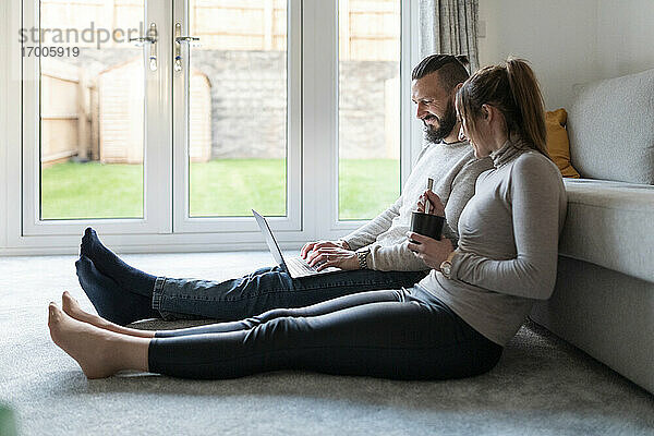 Lächelnde junge Frau trinkt Tee  während ihr Freund zu Hause einen Laptop benutzt