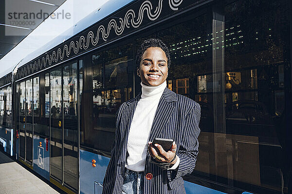 Lächelnde Geschäftsfrau  die ihr Mobiltelefon an einem sonnigen Tag gegen einen Bus hält