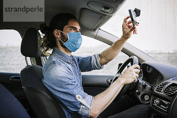 Junger Mann mit Gesichtsschutzmaske beim Einstellen des Spiegels im Auto