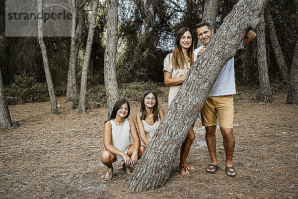 Eltern mit Töchtern stehen hinter einem Baumstamm im Wald