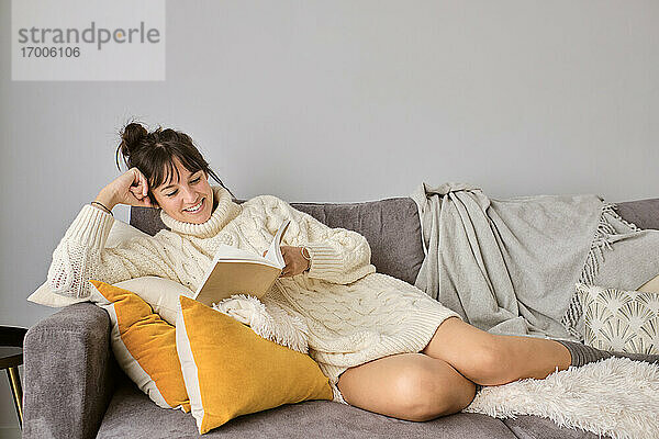 Lächelnde Frau liest ein Buch und entspannt sich auf dem Sofa zu Hause im Winter