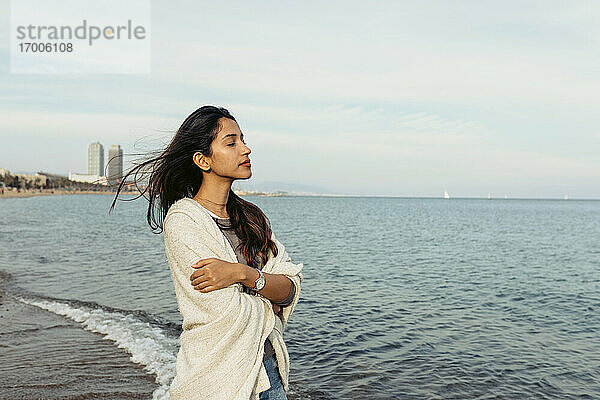 Junge Frau mit verschränkten Armen steht auf dem Wasser gegen den Himmel am Strand
