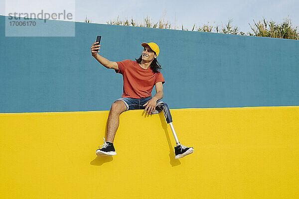 Junger Mann  der ein Selfie macht  während er auf einer mehrfarbigen Wand sitzt