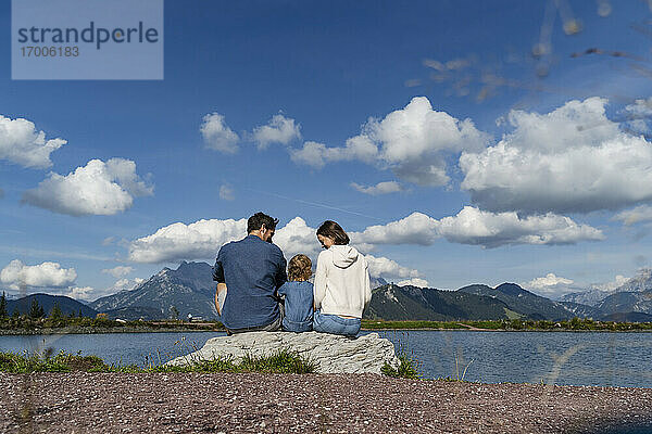 Familie mit kleiner Tochter sitzt gemeinsam auf einem Felsen am Seeufer