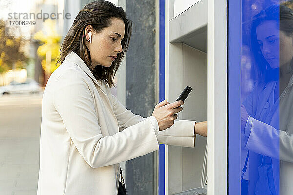 Geschäftsfrau  die ein Smartphone benutzt  während sie Geld vom Geldautomaten in der Stadt abhebt