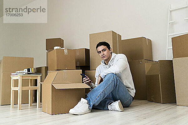 Junger Mann  der ein Mobiltelefon benutzt  während er gegen Kartons in einem neuen Haus sitzt