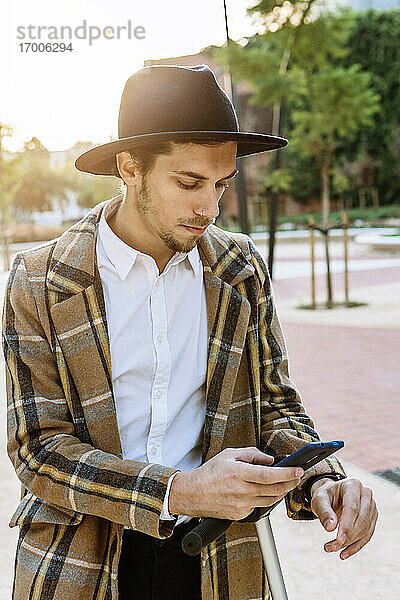Geschäftsmann mit Hut und kariertem Jackett schaut in der Stadt auf sein Smartphone