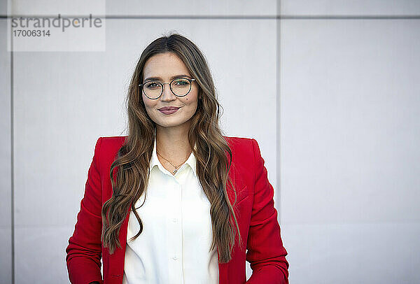Lächelnde Geschäftsfrau mit rotem Blazer an der Wand stehend