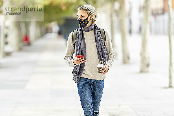 Junger Mann mit Gesichtsmaske  der beim Spaziergang in der Stadt ein Mobiltelefon benutzt