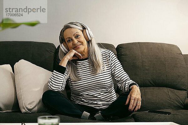 Ältere Frau mit Kopfhörern lächelt  während sie zu Hause mit der Hand am Kinn sitzt