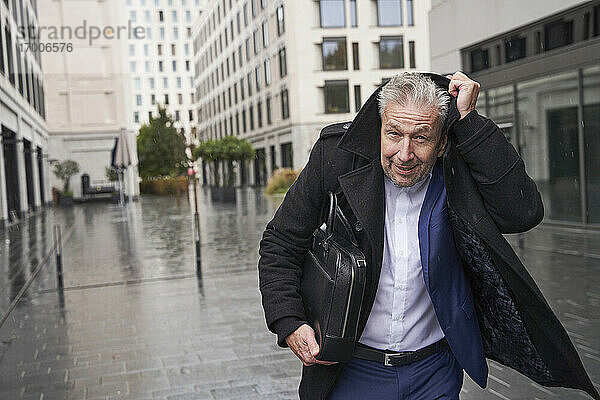 Lächelnder Geschäftsmann mit Aktentasche  der seinen Kopf mit einer Jacke bedeckt  während er im Regen durch die Stadt läuft