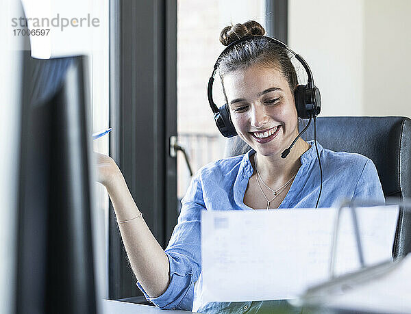 Geschäftsfrau mit Papier  die über Kopfhörer spricht  während sie im Büro sitzt