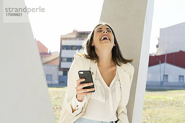 Weibliche Unternehmerin lacht  während sie ein Smartphone in der Stadt an einem sonnigen Tag hält