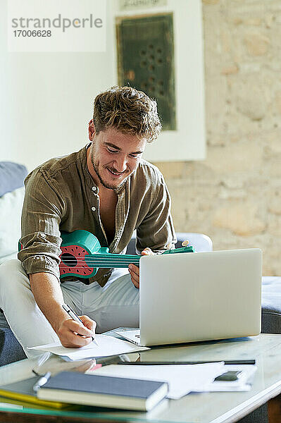 Junger Mann schreibt auf Papier  während er im Wohnzimmer auf einem Laptop online Ukulele lernt
