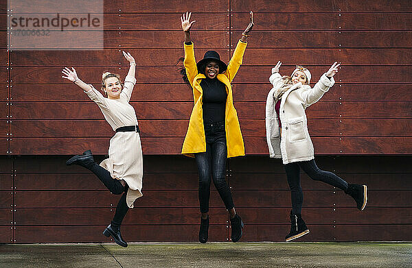Drei glückliche Freundinnen springen vor einer Holzwand