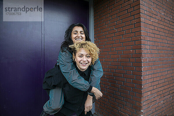 Junge Frau nimmt ihre Freundin vor dem Eingang eines Gebäudes huckepack