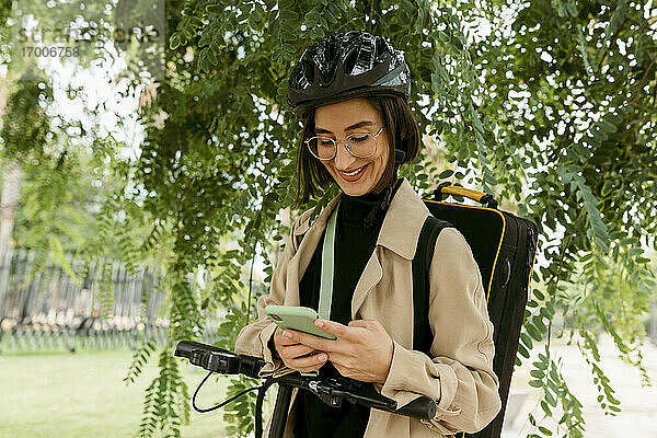 Lächelnde Frau mit Instrumentenkoffer und Elektroroller  die ein Mobiltelefon benutzt  während sie unter einem Baum im Park steht