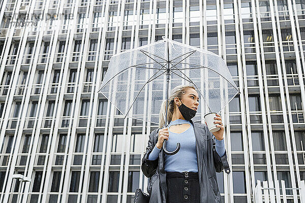 Weibliche Fachkraft mit Regenschirm  die einen Einwegbecher gegen ein Gebäude in der Stadt während COVID-19 hält