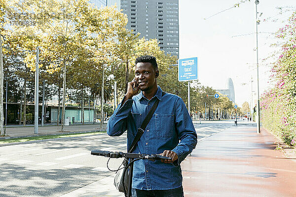 Männlicher Pendler  der mit einem elektrischen Roller auf dem Bürgersteig in der Stadt mit seinem Smartphone telefoniert