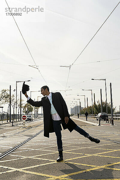 Junger Mann steht auf einem Bein und hält einen Hut zwischen Eisenbahnschienen auf der Straße